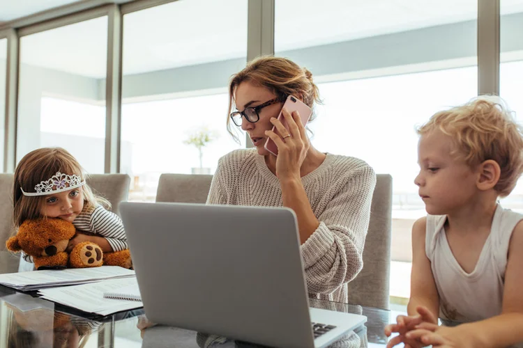 Young woman talking on mobile phone while sitting at tablet with laptop and her kids. Mother working from home with her children sitting by. (jacoblund/Getty Images)