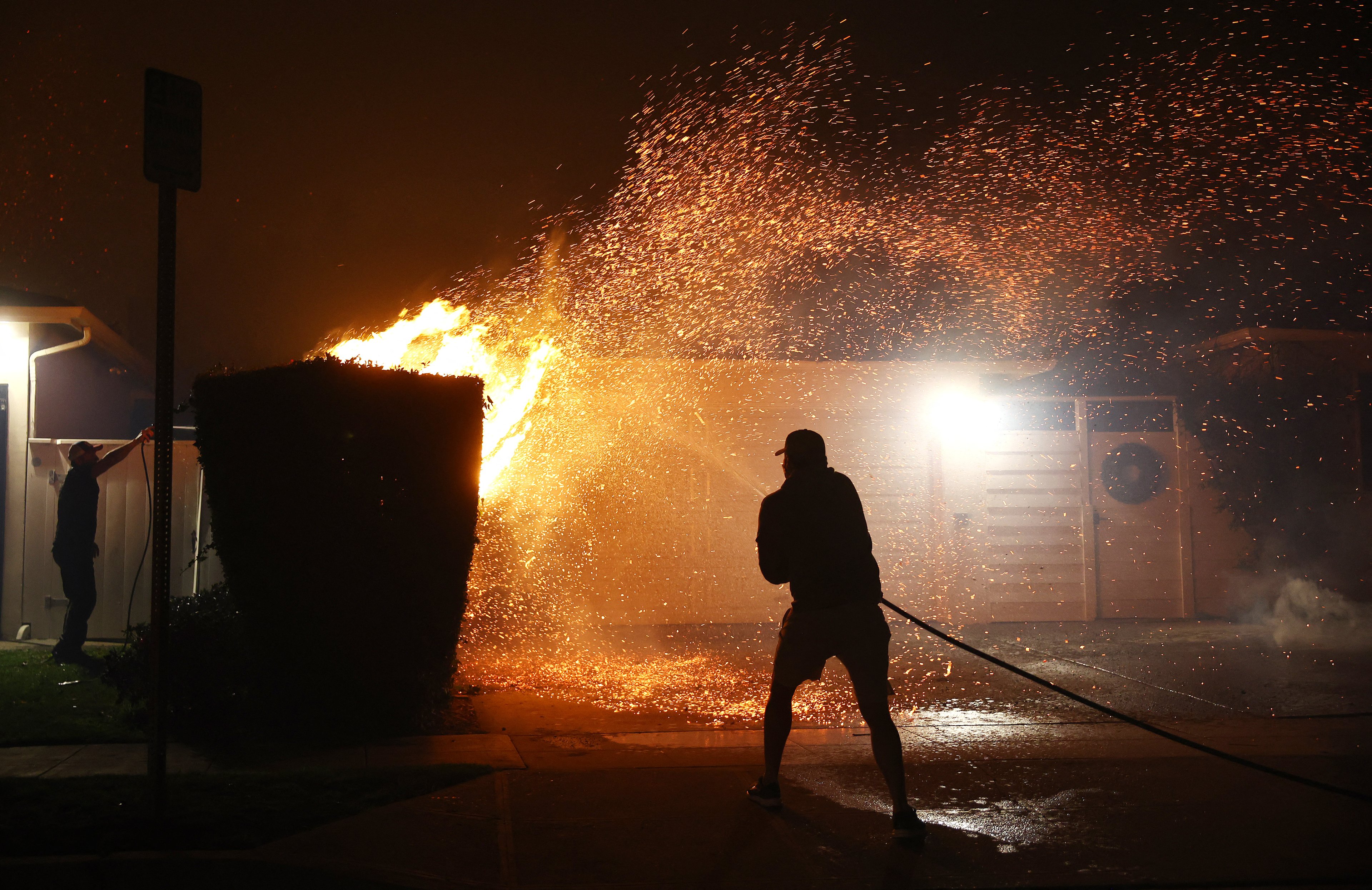 PACIFIC PALISADES, CALIFORNIA - JANUARY 07: A person uses a garden hose to extinguish flames in front of a home as palm trees burn nearby during the Palisades Fire amid a powerful windstorm on January 7, 2025 in Pacific Palisades, California. Fueled by intense Santa Ana Winds, the Palisades Fire has grown to over 2,900 acres and 30,000 people have been ordered to evacuate while a second fire has emerged near Eaton Canyon.   Mario Tama/Getty Images/AFP (Photo by MARIO TAMA / GETTY IMAGES NORTH AMERICA / Getty Images via AFP)