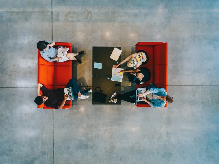 Young people sitting around a table and studying at public library. University students with books and laptop (envato/Divulgação)