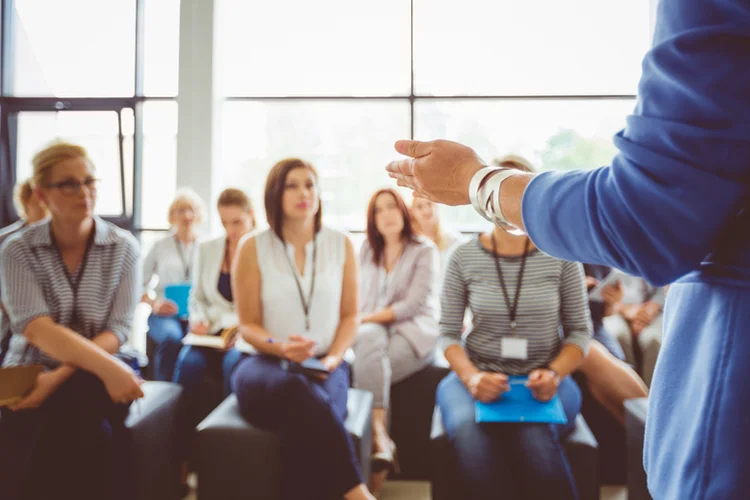 Hand of a trainer addressing group of females sitting in a conference hall. Female hand against defocused group of women attending seminar. (izusek/Getty Images)