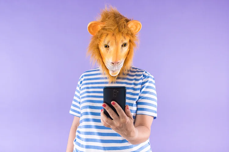 Woman wearing blue striped t-shirt and lion mask, checking her mobile phone, on purple background. Costume, animal, carnival, party and bizarre concept. (Getty Images/DBenitostock)