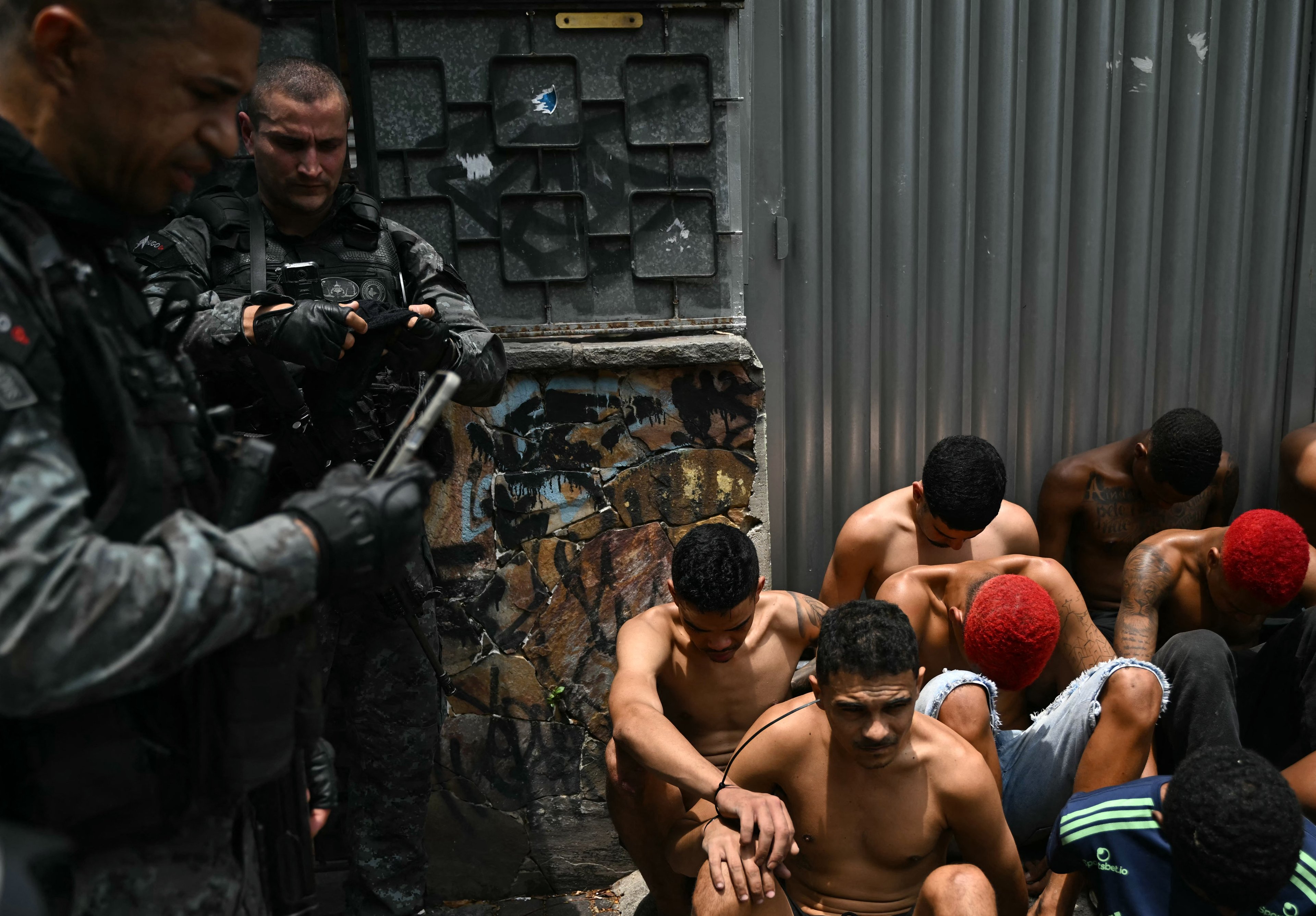 Um policial verifica seu celular ao lado de suspeitos presos durante a Operação Contenção na favela Vila Cruzeiro, no complexo da Penha, no Rio de Janeiro, nesta terça-feira, 28
