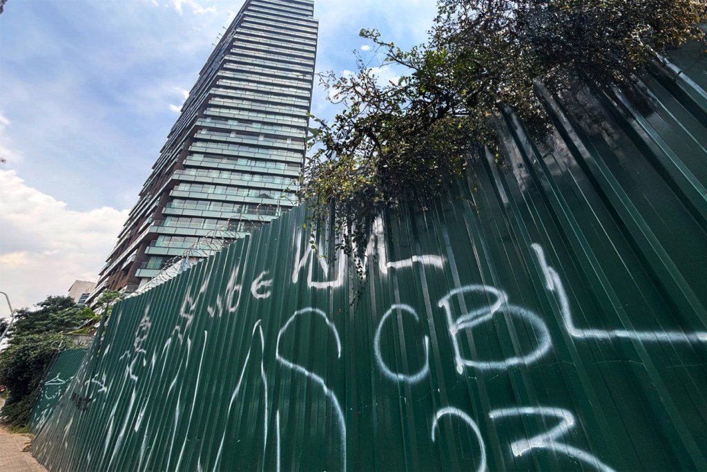 Foto mostra um tapume de metal verde, com pichação em tinta branca. Ao longe tem um prédio alto