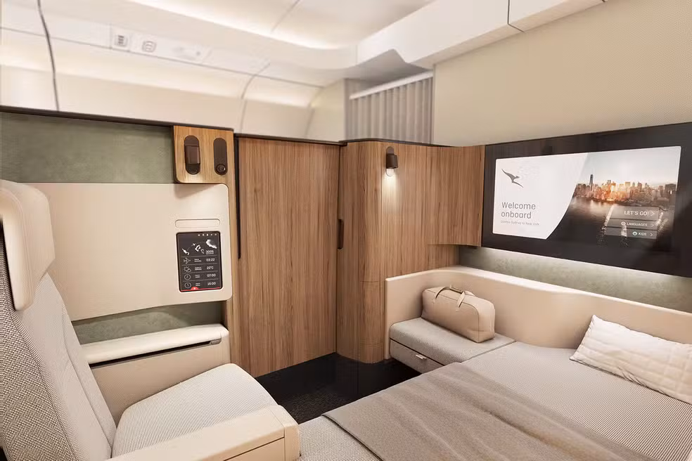 A imagem mostra a cabine da Primeira Classe. É um quarto privativo pequeno, com uma cama compacta, uma poltrona muito confortável, um guarda-roupa, uma mesa e uma televisão de 32 polegadas.
