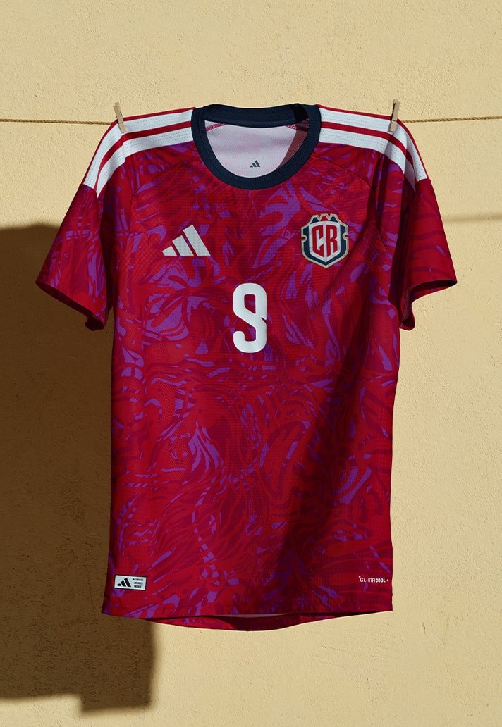 a camisa tem basal  vermelha e traz um gráfico espalhado inspirado nas paisagens bash  país; gola azul‑marinho e o lema “Pura Vida” nas costas complementam o desenho